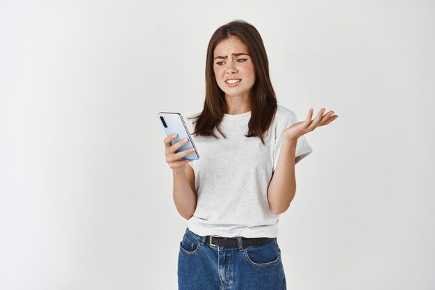 스마트폰에서 어색하고 불편해 보이는 젊은 여성의 이미지, 온라인에서 찡그린 메시지를 받고, 전화 화면을 쳐다보고 어깨를 으쓱하고, 흰색 벽