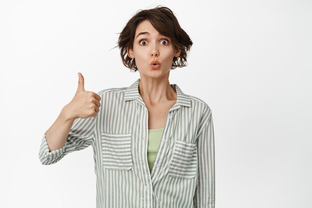 Image of young woman looks impressed, shows thumb up, praise smth very good, like and approve, recommending, leave positive feedback, white background.