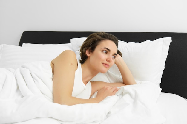 Free photo image of young woman lying in bed resting at home under white linen sheets looking with pleased