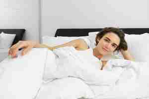 Free photo image of young woman lying in bed resting at home under white linen sheets looking with pleased