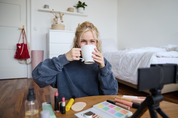 Foto gratuita immagine di una giovane vlogger di trucco seduta in camera digitale in camera da letto che beve tè e parla