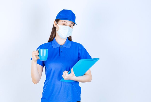 Image of young woman in medical mask holding blue folder and cup. 