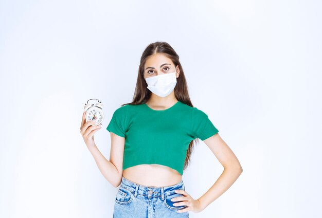 Image of young woman in medical mask posing with alarm clock.