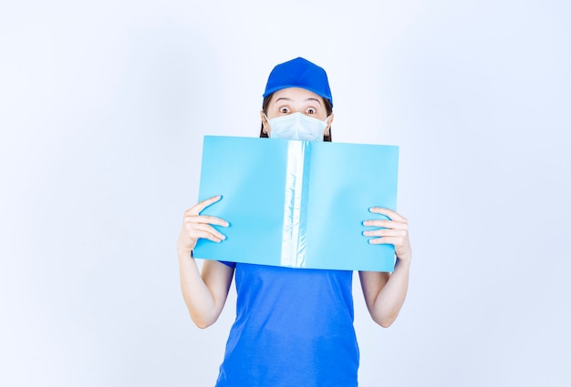 Image of young woman in medical mask posing with blue folder over white wall. 