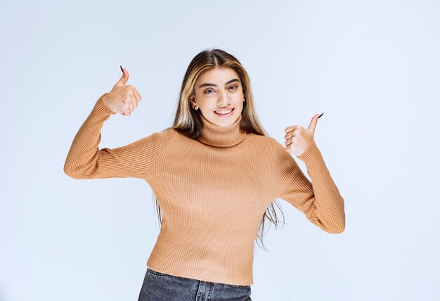Изображение модели молодой женщины в коричневом свитере стоя и показывает палец вверх.