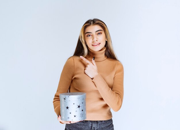 Image of a young woman model in brown sweater with a gift box pointing away.