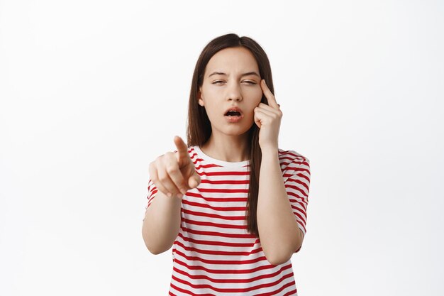 Image of young woman pointing finger, squinting eyes without glasses, cant see, trying to read smth nog wearing eyeglasses, standing in striped t-shirt against white background