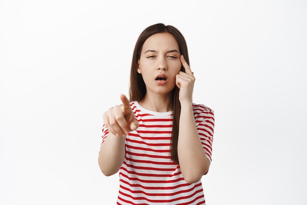 Image of young woman pointing finger, squinting eyes without glasses, cant see, trying to read smth nog wearing eyeglasses, standing in striped t-shirt against white background