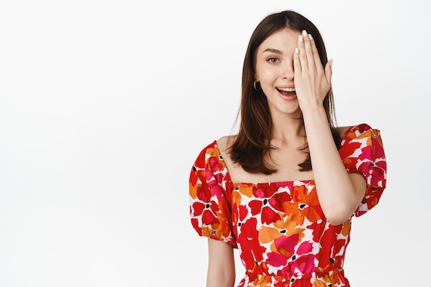 Image of young woman in summer dress cover half of face looking with on eye and smiling at camera white background