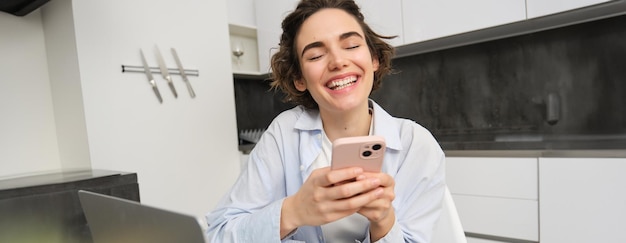 Free photo image of young woman using her smartphone at home girl sits with mobile phone in kitchen and smiles