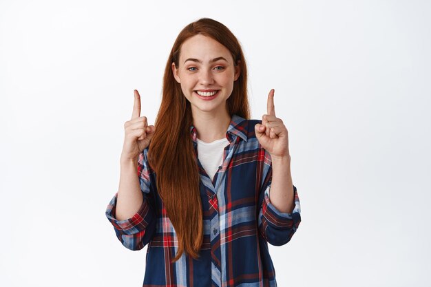 Image of young woman with natural face and long red hair, pointing fingers up, smiling white teeth, give information, advertising, standing over white background.