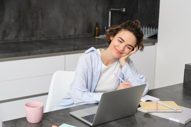 Image of young woman works on laptop at home productive girl studies in her kitchen on computer list