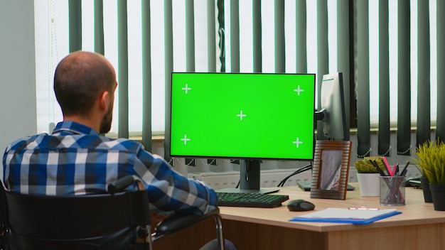 화상 회의를 위해 크로마 키가 있는 컴퓨터를 사용하여 휠체어에 고정된 사업가입니다. 녹색 화면, 모형, 원격 동료와 대화하는 키가 있는 PC를 보고 있는 장애인 프리랜서