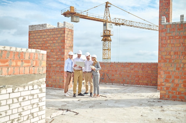 Important information. Three men in white shirt and protective helmet and woman in business suit with drawings looking into laptop standing in building under construction