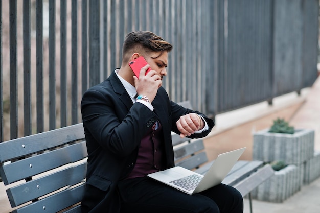 Un incontro importante elegante uomo d'affari indiano in abbigliamento formale seduto su una panchina con il laptop e guardando gli orologi mentre parla al telefono