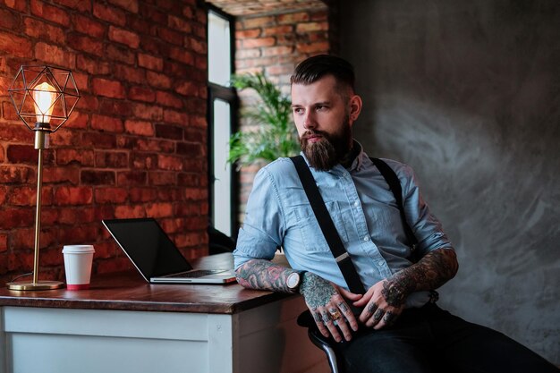 인상적인 수염 난 남자가 그의 사무실에 있는 테이블에 기대어 있습니다. 그는 셔츠와 멜빵을 입고 있습니다. 그는 팔에 문신이 있습니다. 배경에는 노트북과 커피 한 잔이 있습니다.