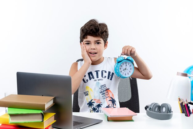 Impreased little schoolboy sitting at desk with school tools holding alarm clock and putting hand on cheek isolated on white background