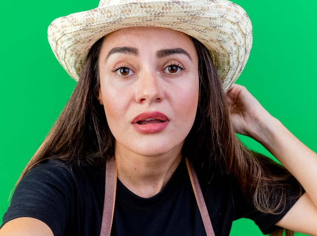 Impresed beautiful gardener girl in uniform wearing gardening hat holding camera isolated on green background