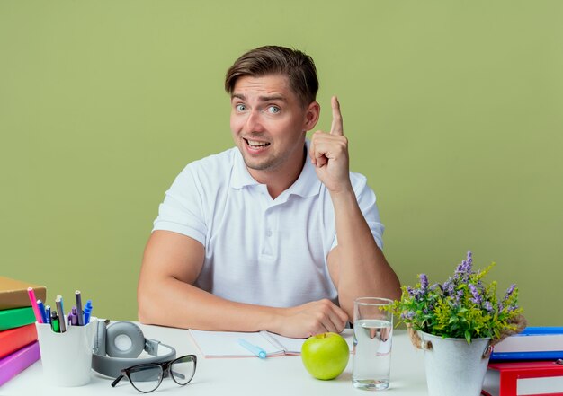 impresed young handsome male student sitting at desk with school tools points at up on olive green