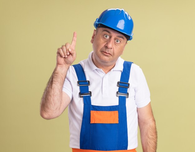 Impressed adult builder man in uniform points up isolated on olive green wall