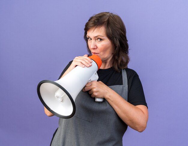 Impressed adult caucasian female barber in uniform holding loud speaker and looking at camera isolated on purple background with copy space