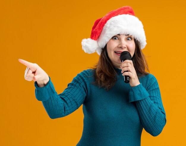 impressed adult caucasian woman with santa hat holding mic and pointing at side isolated on orange background with copy space