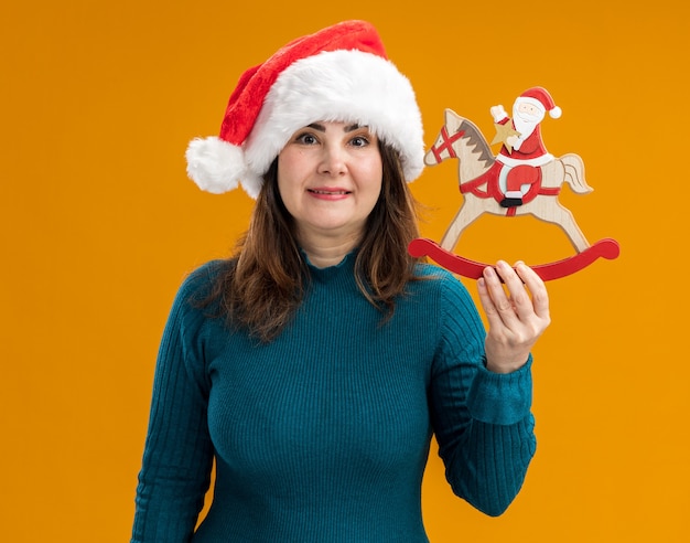 Impressed adult caucasian woman with santa hat holding santa on rocking horse decoration isolated on orange background with copy space