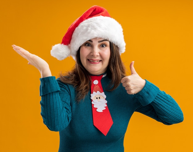 Impressed adult caucasian woman with santa hat and santa tie thumbs up and holds hand open isolated on orange background with copy space
