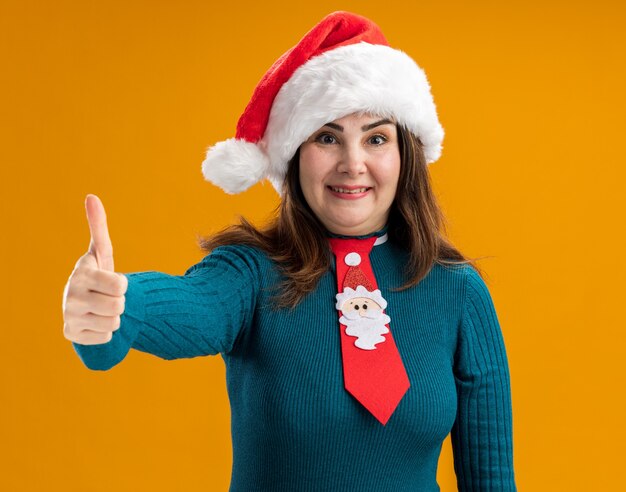 Impressed adult caucasian woman with santa hat and santa tie thumbs up isolated on orange background with copy space