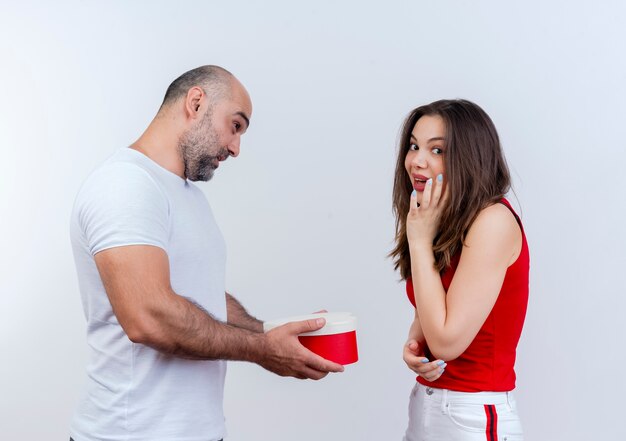 Impressed adult couple man holding heart-shaped gift box looking at woman and woman touching face and looking isolated