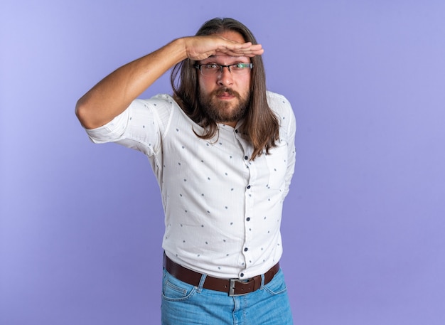 Impressed adult handsome man wearing glasses keeping hand behind back and another hand on forehead looking at camera into distance isolated on purple wall with copy space