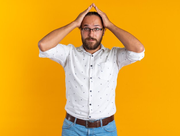 Impressed adult handsome man wearing glasses keeping hands on head looking at camera isolated on orange wall