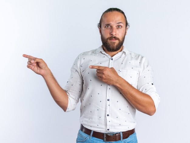 Impressed adult handsome man wearing glasses looking at camera pointing at side isolated on white wall