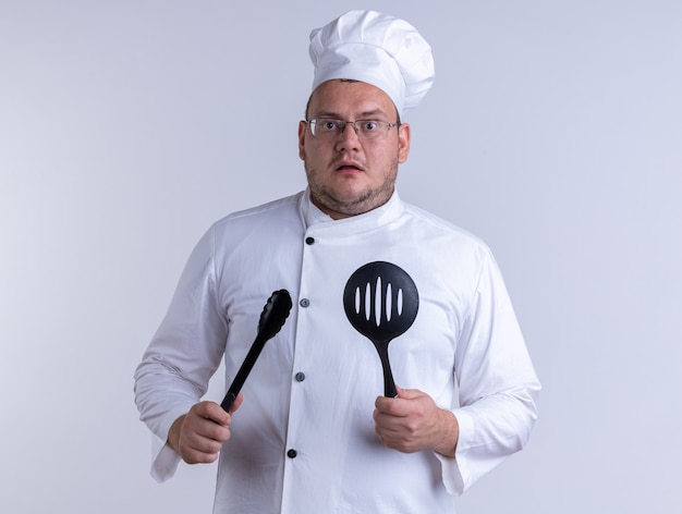 Бесплатное фото Впечатленный взрослый мужчина-повар в униформе шеф-повара и в очках держит щипцы и шумовку, глядя вперед, изолированную на белой стене