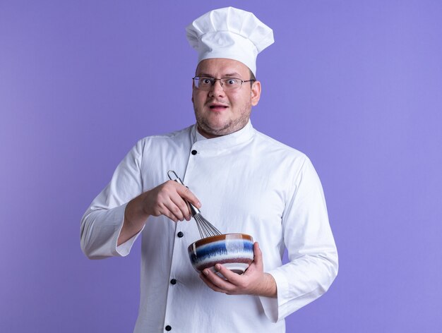 Impressed adult male cook wearing chef uniform and glasses holding bowl and whisk looking at camera isolated on purple background