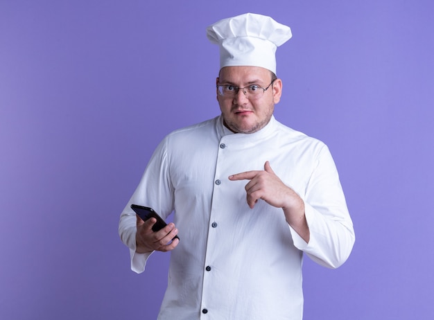 impressed adult male cook wearing chef uniform and glasses holding and pointing at mobile phone looking at front isolated on purple wall with copy space