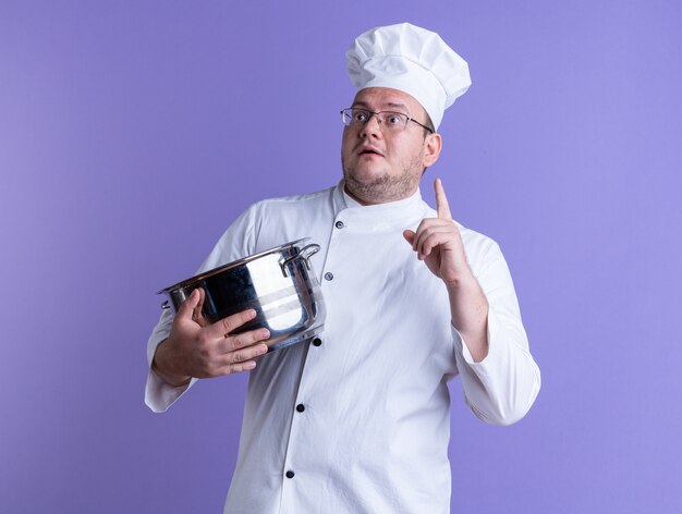 impressed adult male cook wearing chef uniform and glasses holding pot looking at side pointing up isolated on purple wall