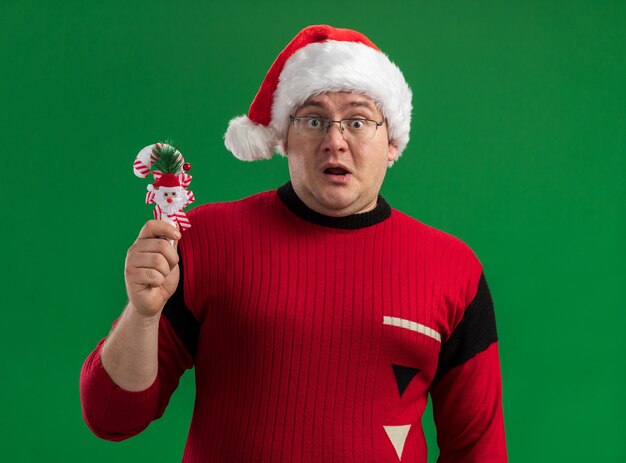 안경과 녹색 배경에 고립 된 카메라를보고 사탕 지팡이 장식을 들고 산타 모자를 쓰고 감동 된 성인 남자