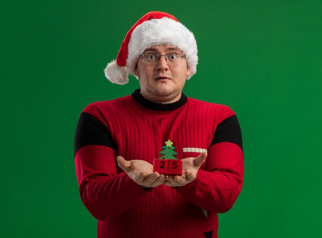 Impressed adult man wearing glasses and santa hat holding christmas tree toy with date  looking at camera isolated on green background
