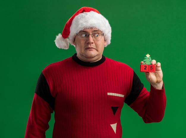 Free photo impressed adult man wearing glasses and santa hat holding and looking at christmas tree toy with date  isolated on green background