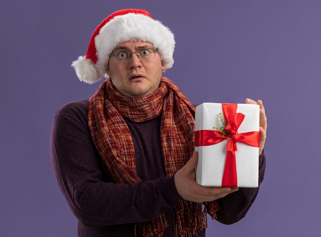 Impressed adult man wearing glasses and santa hat with scarf around neck holding gift package  isolated on purple wall