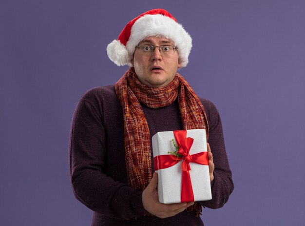 impressed adult man wearing glasses and santa hat with scarf around neck holding gift package looking at camera isolated on purple background