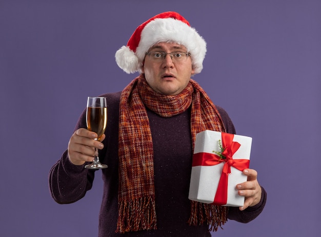 보라색 벽에 고립 된 샴페인과 선물 패키지의 유리를 들고 목에 스카프와 안경과 산타 모자를 쓰고 감동 성인 남자