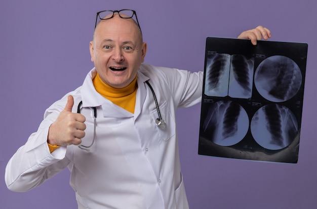 Impressed adult man with glasses in doctor uniform with stethoscope holding x-ray result and thumbing up