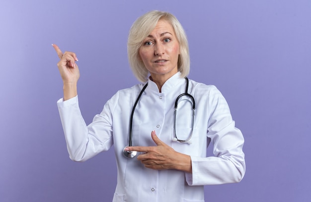 Бесплатное фото Впечатленная взрослая славянская женщина-врач в медицинском халате со стетоскопом, указывающим на сторону, изолированную на фиолетовом фоне с копией пространства