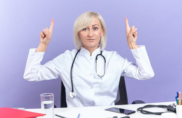 Free photo impressed adult slavic female doctor in medical robe with stethoscope sitting at desk with office tools pointing up isolated on purple wall with copy space