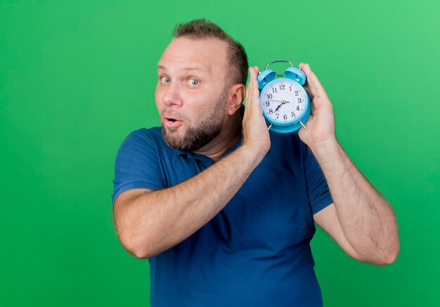 Impressed adult slavic man holding alarm clock looking  isolated on green wall with copy space