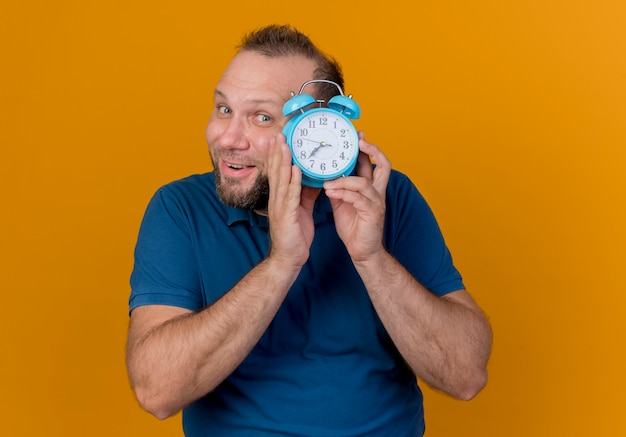 Impressed adult slavic man holding alarm clock near head and looking from behind it