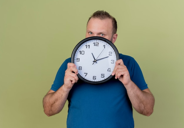 Impressed adult slavic man holding clock and looking from behind it 