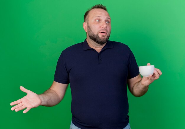 Impressed adult slavic man holding cup showing empty hand looking at side isolated on green wall